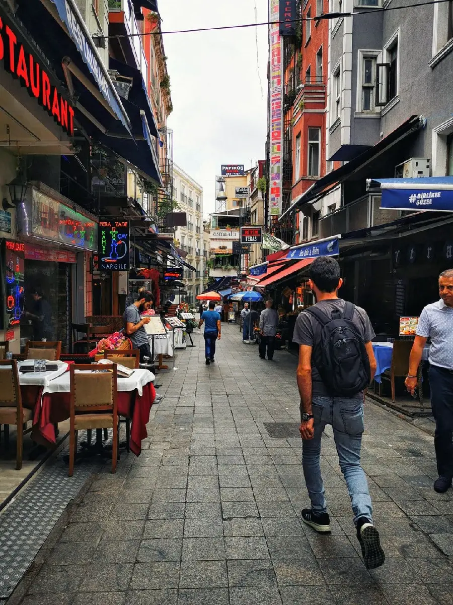 A bright and sunny image From a narrow city road with restaurants on both sides and pedestrians