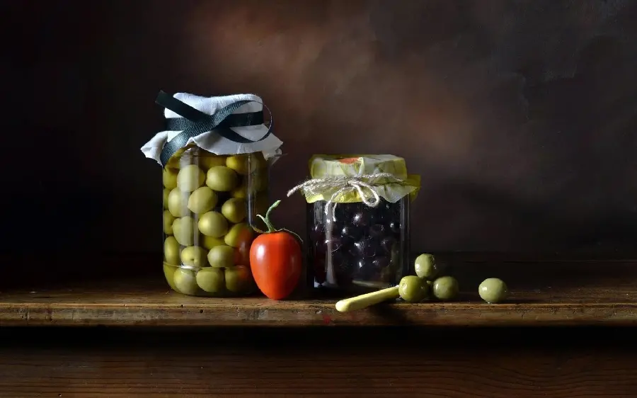 A luxurious image of two beautiful bottles of black and green olives with a tomato between them on a brown background