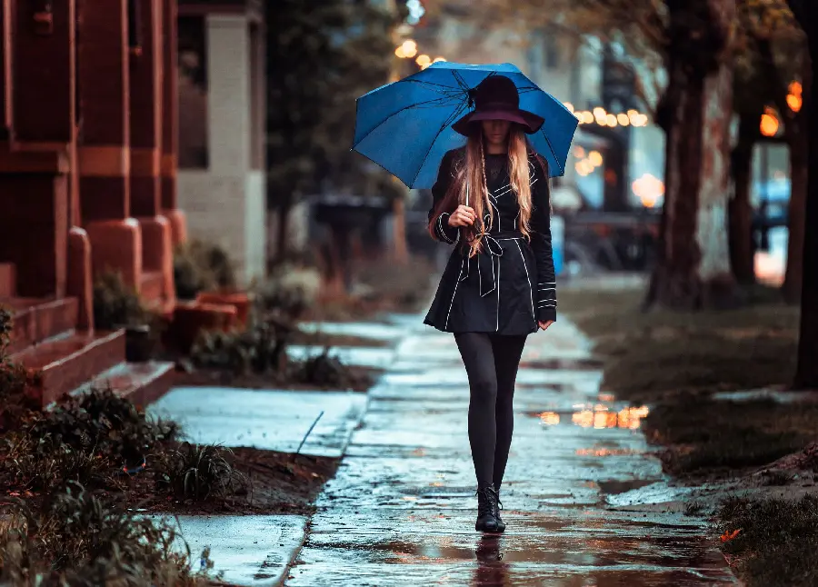 A lovely picture of a classy girl with a beautiful hat on her head and a blue umbrella in real rain and next to beautiful houses