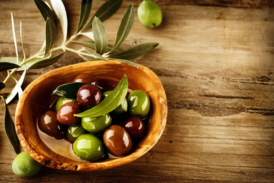 Photo of a wooden bowl of olives on a wooden board with leaves next to it
