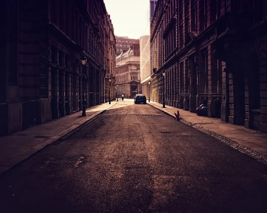 Download a nostalgic and dimly lit photo with tall, old buildings and a passerby on an urban road