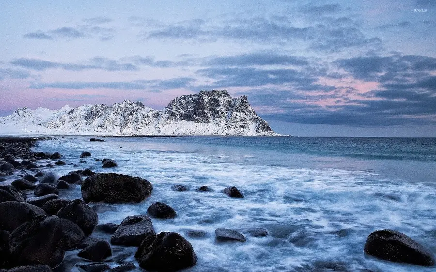 Download a lovely fantasy photo of the calm ocean and black rocks next to it and snowy mountains in high quality