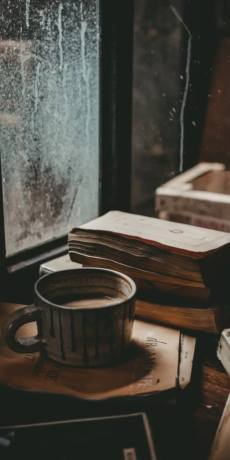 Sad profile of a coffee cup next to a book and a rainy window