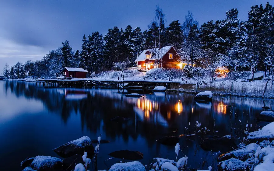 Home A snowy landscape with lights on the edge of a Scandinavian lake	